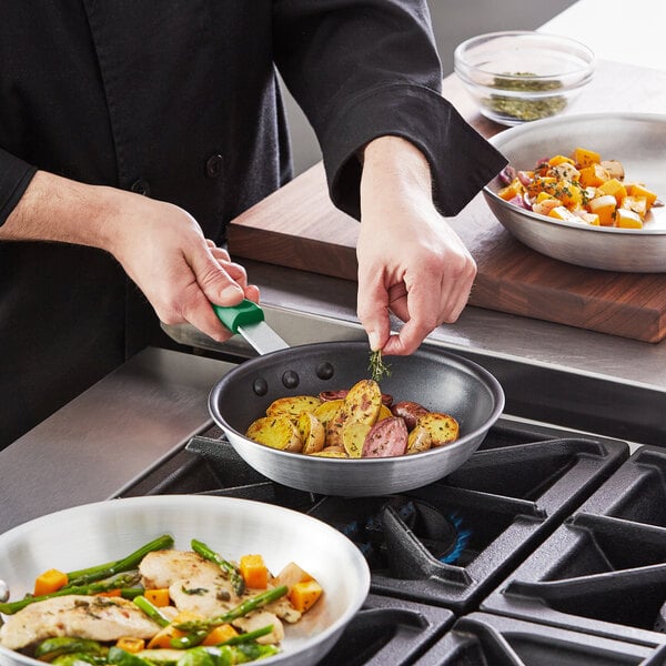 A person using a Choice aluminum non-stick fry pan with a green silicone handle to cook food on a stove.
