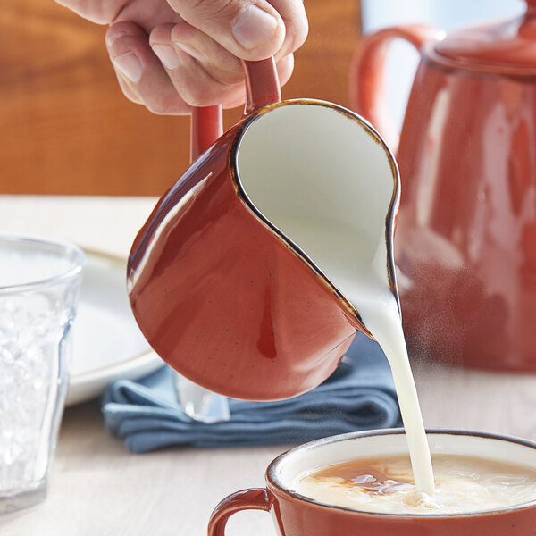 A person pouring milk from a red pitcher into a white cup.