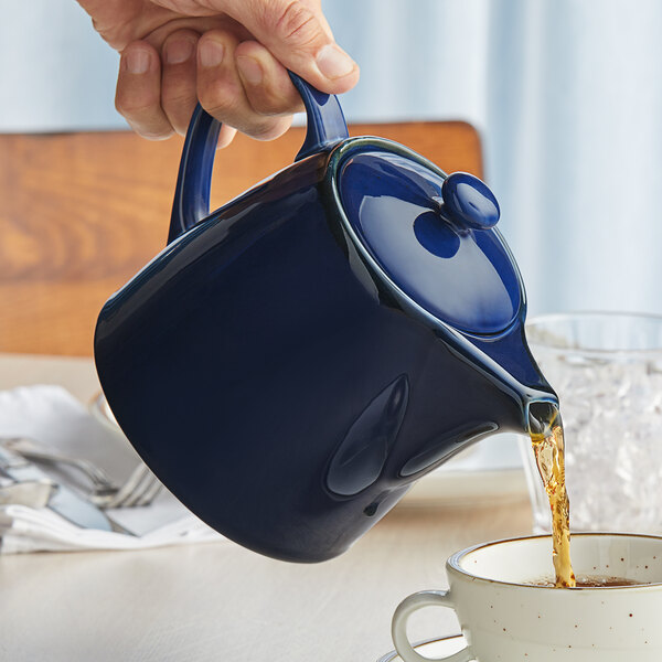 A hand pouring tea into a blue Acopa Keystone stoneware teapot.