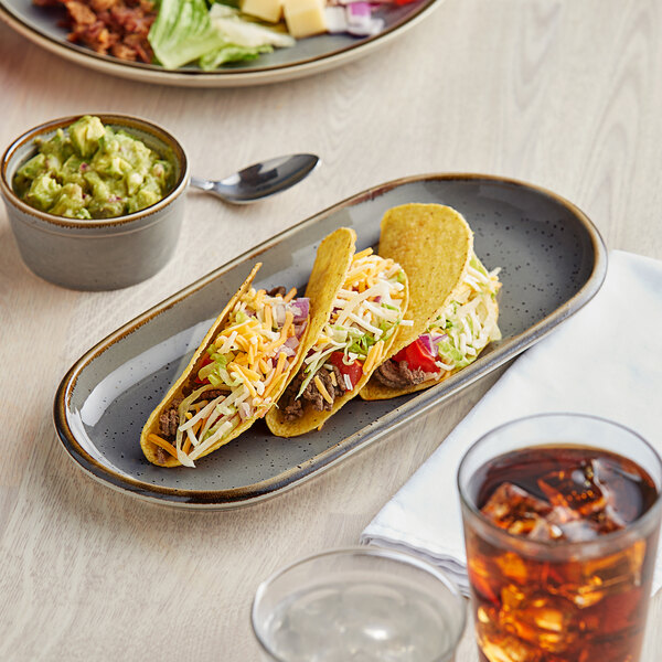 An Acopa granite gray stoneware oblong coupe platter with tacos, a bowl of guacamole, and a drink on a table.