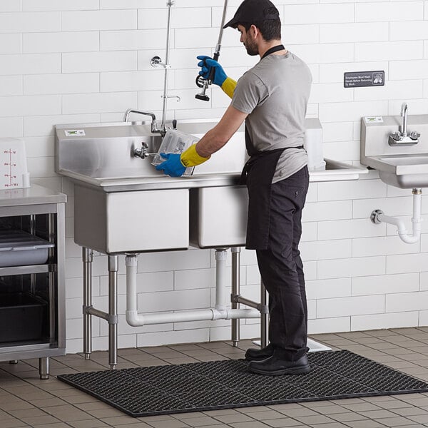 A man in yellow gloves and a blue shirt washing a Regency stainless steel commercial sink.