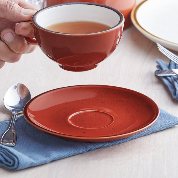 A hand holding a red and white Acopa stoneware cup of tea over a Sedona orange stoneware saucer.
