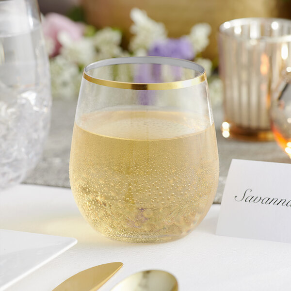 A Visions clear plastic stemless wine glass with a gold rim filled with a clear liquid on a table.