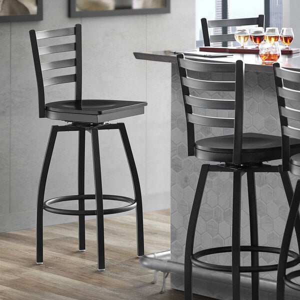 A Lancaster Table & Seating black metal ladder back swivel bar stool with a black wood seat next to a table.