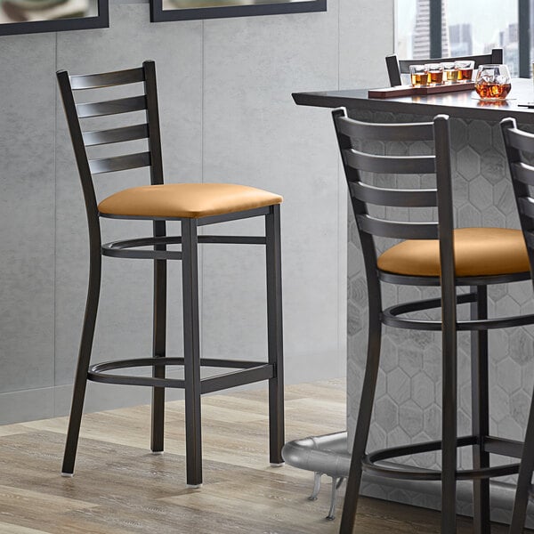 A Lancaster Table & Seating copper ladder back bar stool with a light brown cushion next to a table.