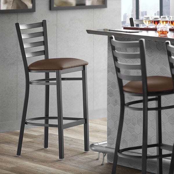 A Lancaster Table & Seating black ladder back bar stool with dark brown vinyl padded seat.