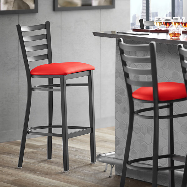 A Lancaster Table & Seating black ladder back bar stool with red vinyl padded seat next to a table.