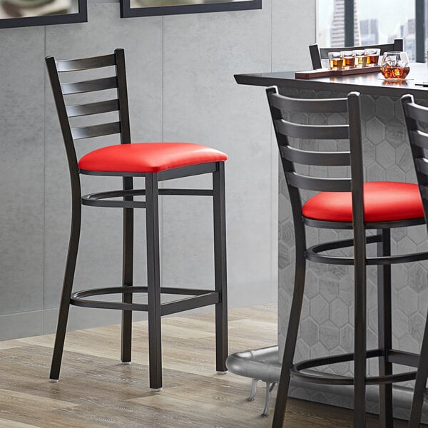 A Lancaster Table & Seating distressed copper ladder back bar stool with a red vinyl padded seat next to a table.