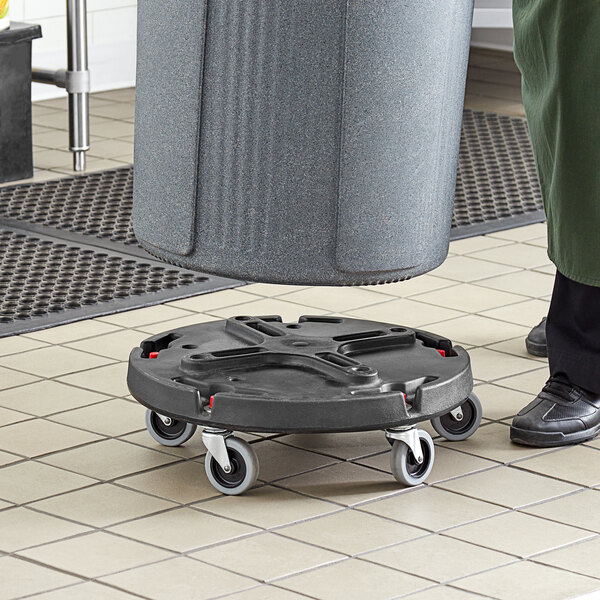 A person using a black Toter caster dolly to move a trash can.