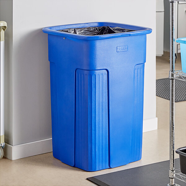 A blue Toter Slimline plastic trash can next to a wall.