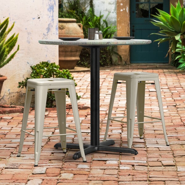 A Lancaster Table & Seating Excalibur outdoor table base with two stools on a brick patio.