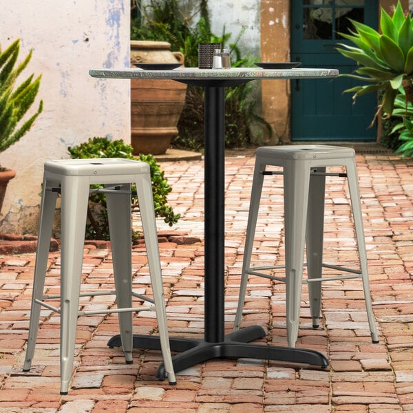 A black Lancaster Table & Seating outdoor table base with white bar height stools on a brick patio.