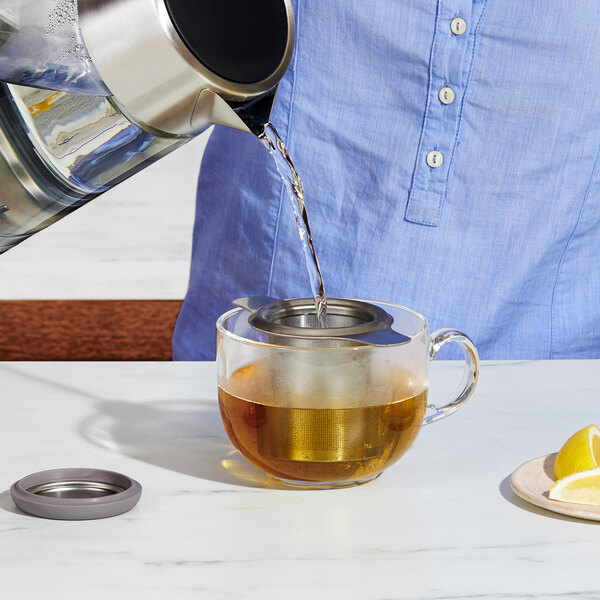A person using an OXO stainless steel tea infuser to pour tea into a glass teapot.