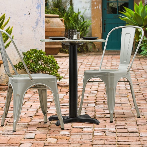 A black Lancaster Table & Seating Excalibur outdoor table base with a black pole on a table with two chairs on a brick patio.