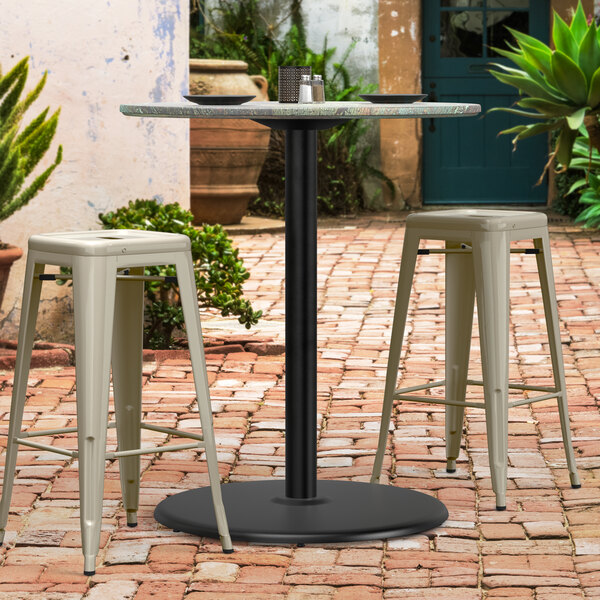 A black Lancaster Table & Seating outdoor table base with white stools on a brick patio.