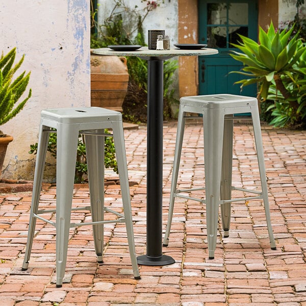 A Lancaster Table & Seating Excalibur black bolt down table base with a 3" bar height column on a brick patio with white stools.