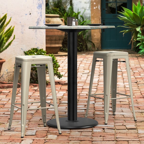 A Lancaster Table & Seating Excalibur black outdoor table base with two white bar stools on a brick patio.