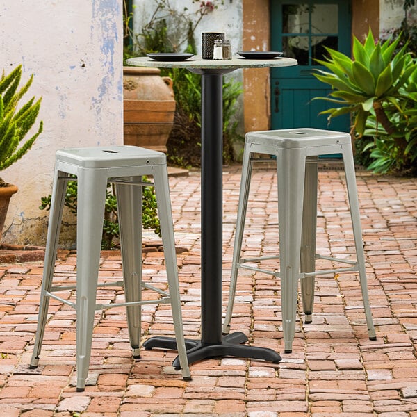 A Lancaster Table & Seating black outdoor table base with bar height pole and two white stools on a brick patio.