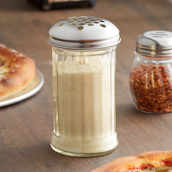 A glass cheese shaker with a silver metal lid on a table with food.