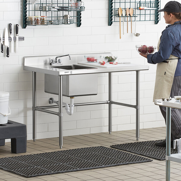 A stainless steel work table with a sink, cross brace support, and a person preparing food beside it in a commercial kitchen setting.