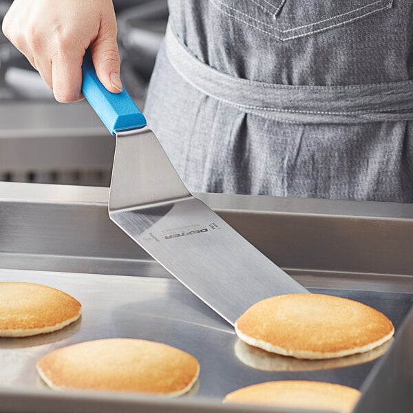 A hand holding a blue Dexter-Russell cake turner over pancakes on a pan.