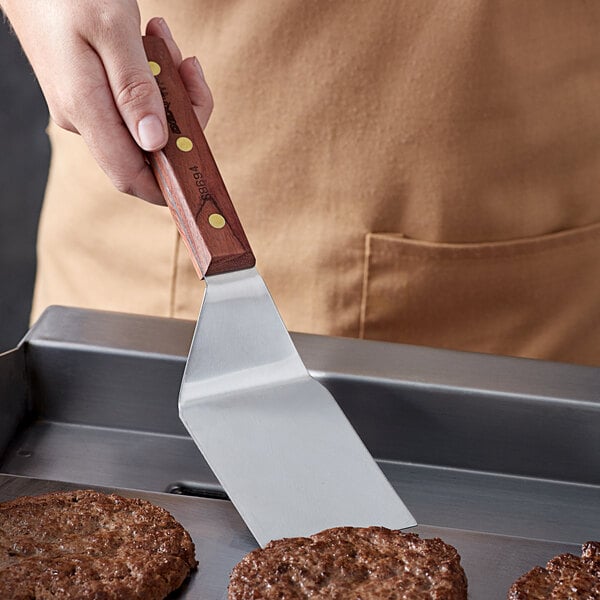A person holding a Dexter-Russell rosewood hamburger turner over a hamburger patty cooking on a pan.