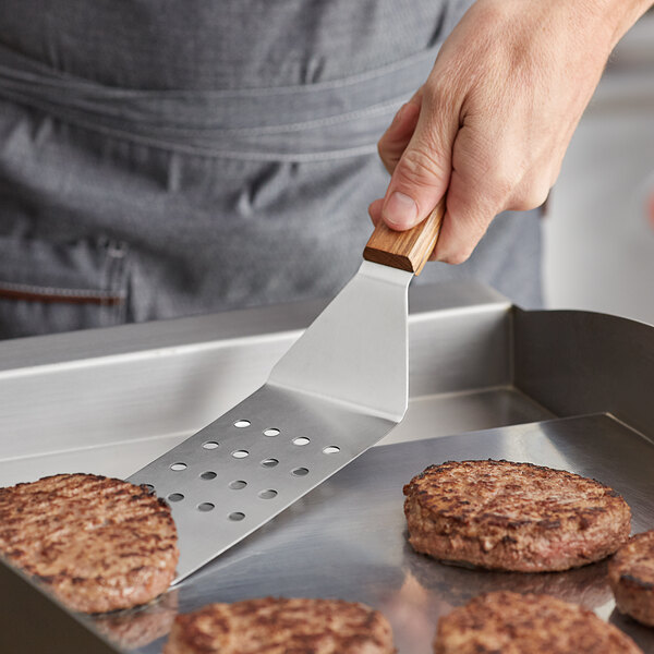 A person using a Vollrath perforated stainless steel hamburger turner to cook burgers on a grill.