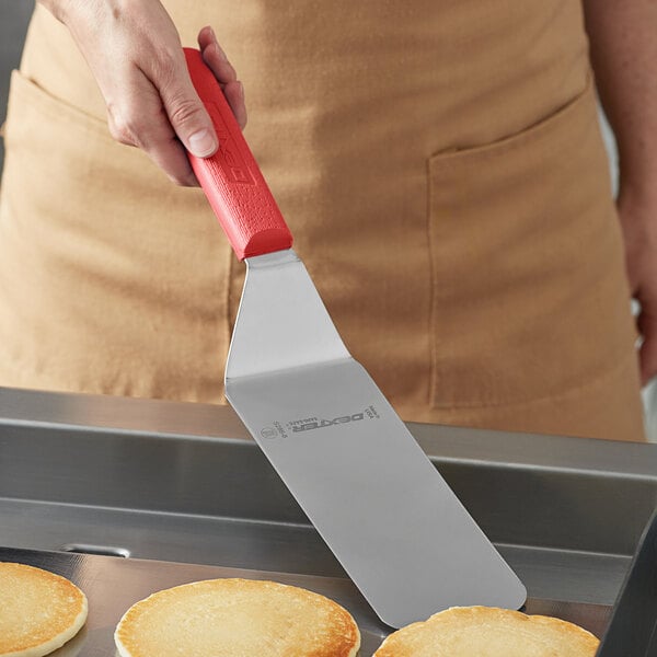 A person holding a Dexter-Russell solid turner with a red handle over a pancake on a plate.