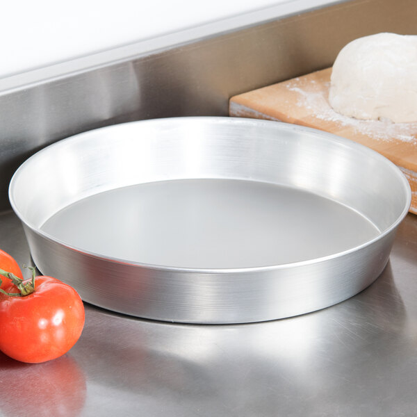 An American Metalcraft tin-plated steel deep dish pizza pan with a tomato next to dough and a cutting board.