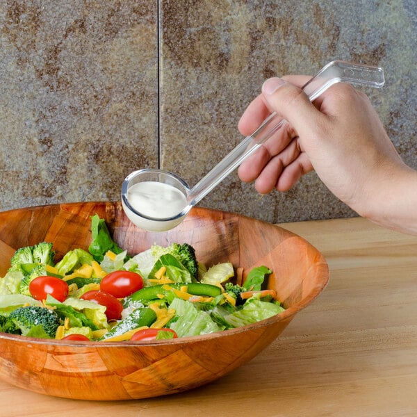 A hand holding a Cambro clear plastic ladle pouring dressing into a bowl of salad.