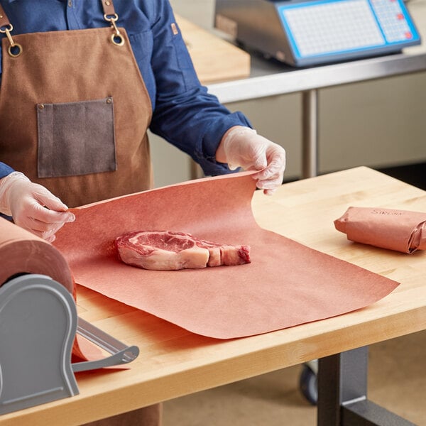 A person in a brown apron in a butcher shop using Choice pink butcher paper to wrap meat on a table.