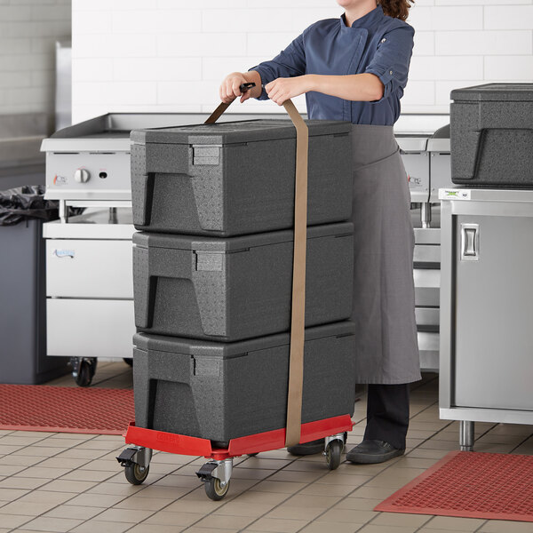 A woman using a grey Cambro Cam GoBox Pan Carrier to push a stack of black plastic boxes on a cart.