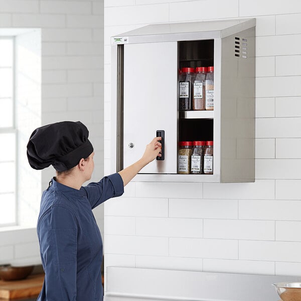 A woman in a chef hat opening a Regency stainless steel wall cabinet with sliding doors.