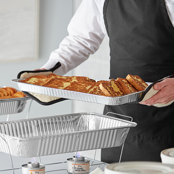 A person holding a Choice shallow foil steam table pan of food on a table at a catering event.
