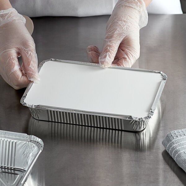 A person in gloves holding a white rectangular shallow foil container with a clear plastic lid.