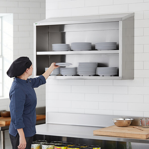A woman wearing a chef hat putting plates on a Regency stainless steel wall shelf.