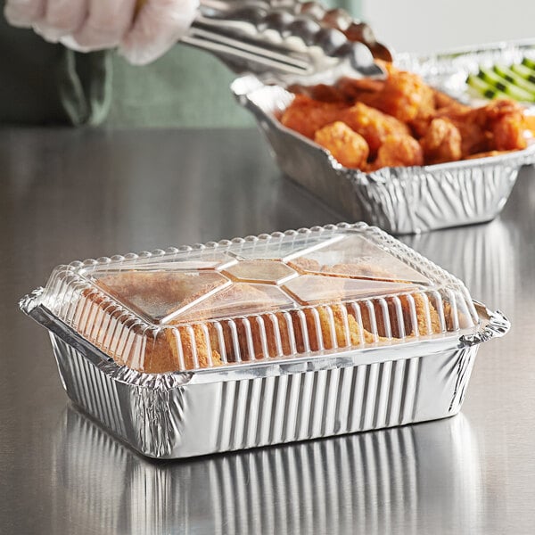 A close-up of a hand holding a fork and knife over chicken in a Choice oblong foil container with a dome lid.