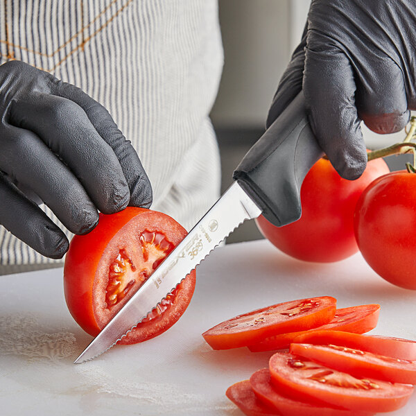 A person in black gloves uses a Dexter-Russell scalloped utility knife to cut a tomato on a counter.