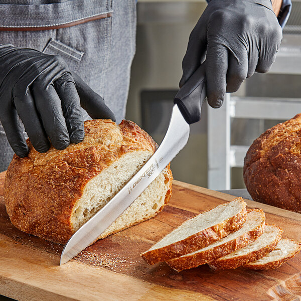 A person wearing black gloves using a Dexter-Russell scalloped bread knife to cut a loaf of bread on a cutting board.