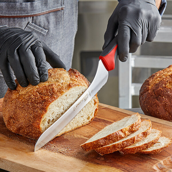 A person using a Dexter-Russell red-handled bread knife to cut a loaf of bread on a cutting board.