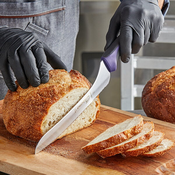 A person wearing black gloves uses a Dexter-Russell purple bread knife to cut a loaf of bread on a cutting board.