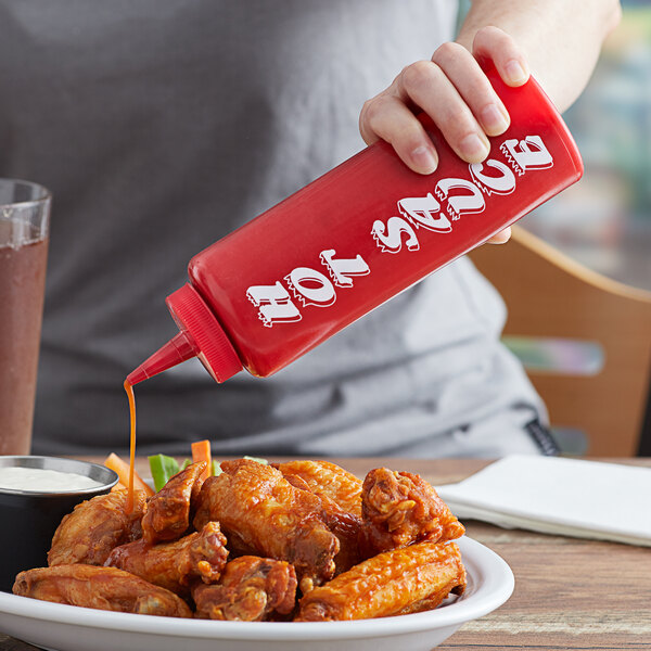A person using an AllPoints red squeeze bottle to pour hot sauce on a plate of chicken wings.