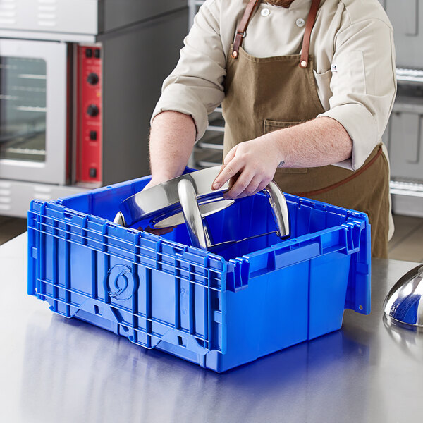A man in an apron holding a blue Orbis Stack-N-Nest Flipak tote box with metal objects inside.