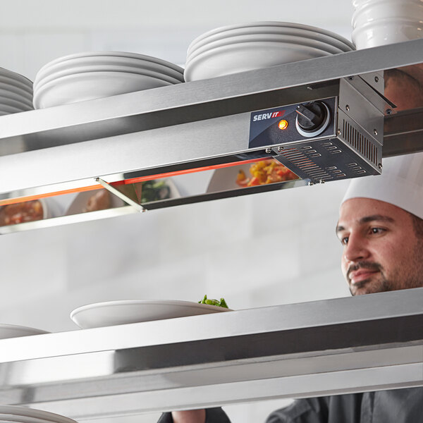 A chef in a professional kitchen looks at a shelf of white plates above a ServIt strip warmer.