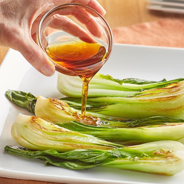 A hand pouring brown Lee Kum Kee Pure Sesame Oil onto a plate of green vegetables.