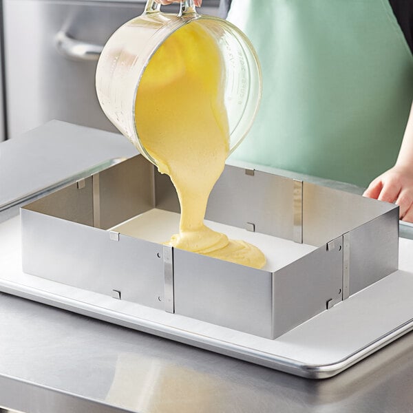 A person in a green apron pouring yellow liquid into a metal rectangular cake ring.