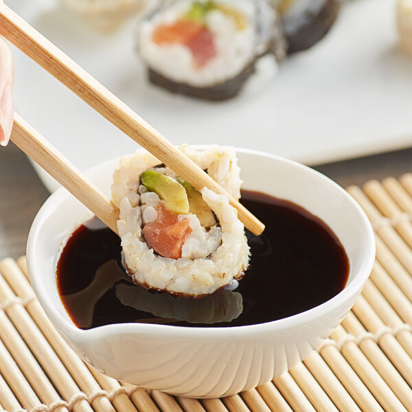 A hand using chopsticks to dip a sushi roll into a bowl of soy sauce.