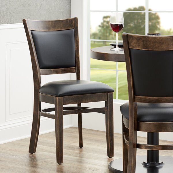 A pair of Lancaster Table & Seating Sofia wood chairs with black vinyl seats at a table with a glass of wine.