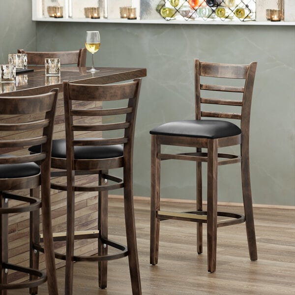 A Lancaster Table & Seating wood bar stool with a black vinyl seat.