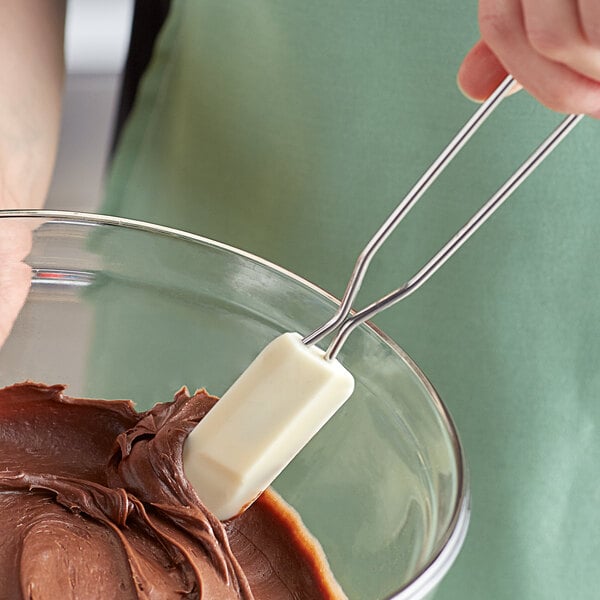 A person using a white Linden Sweden silicone spatula with a metal handle to mix chocolate frosting in a bowl.